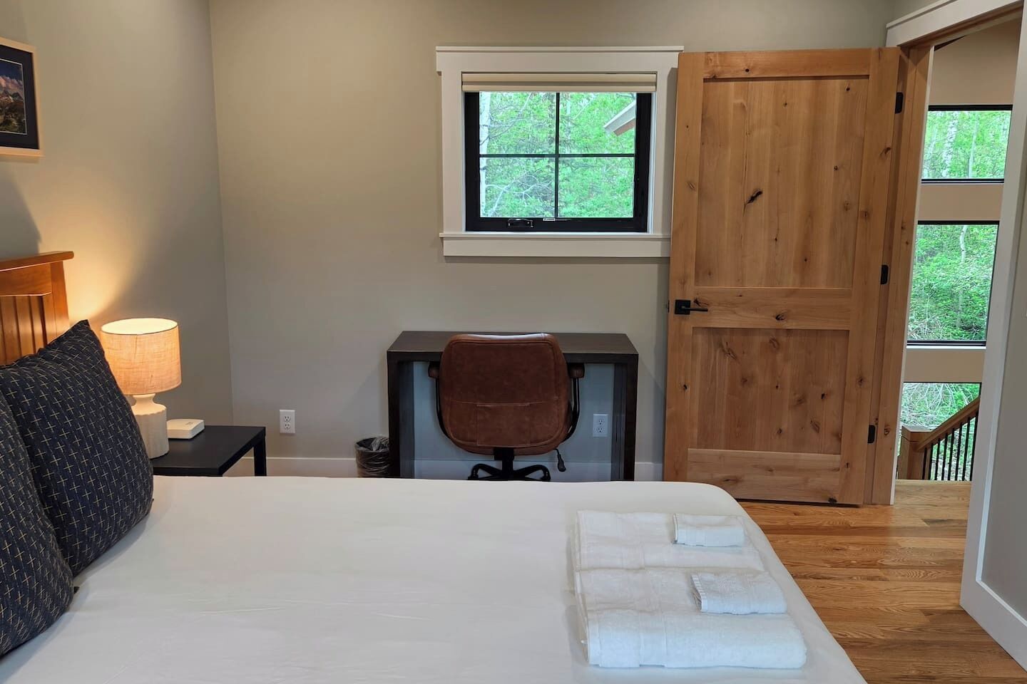 A bedroom with a bed, a desk, a chair, a window, and a door leading to a balcony.