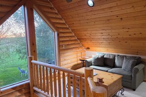 Ein gemütliches Blockhaussinnenzimmer mit Holzbalkendecke, einer Couch, einem Tisch und Blick auf eine grüne Landschaft.