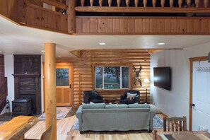 A log cabin interior with a sofa, chairs, a fireplace, a TV, and a window with a view of trees. - Wild Cat Lodge with Fenced Yard and Teton View (Driggs)