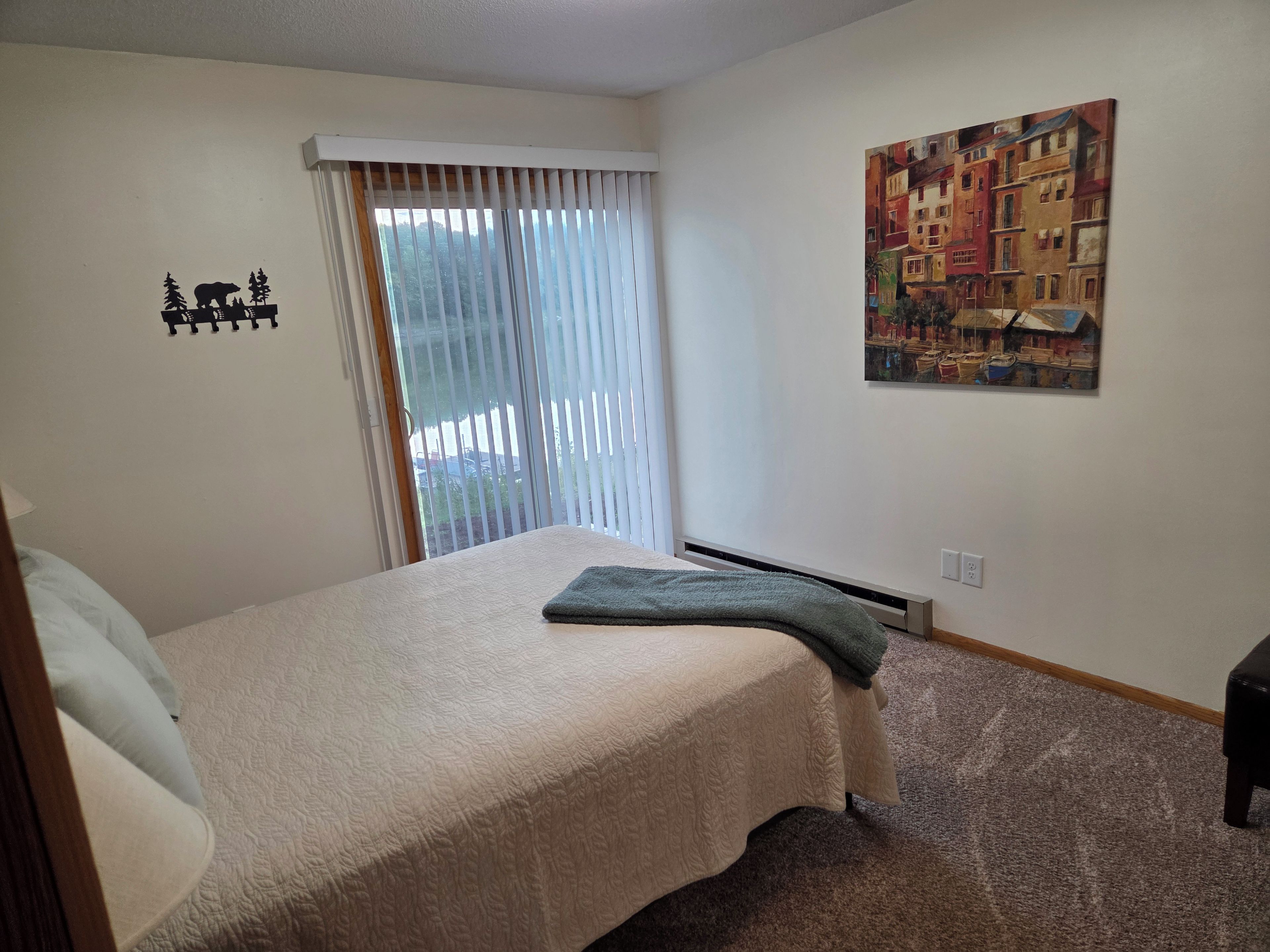 Une chambre à coucher avec un lit, une fenêtre avec des stores, un tableau au mur et un sol recouvert d’une moquette.