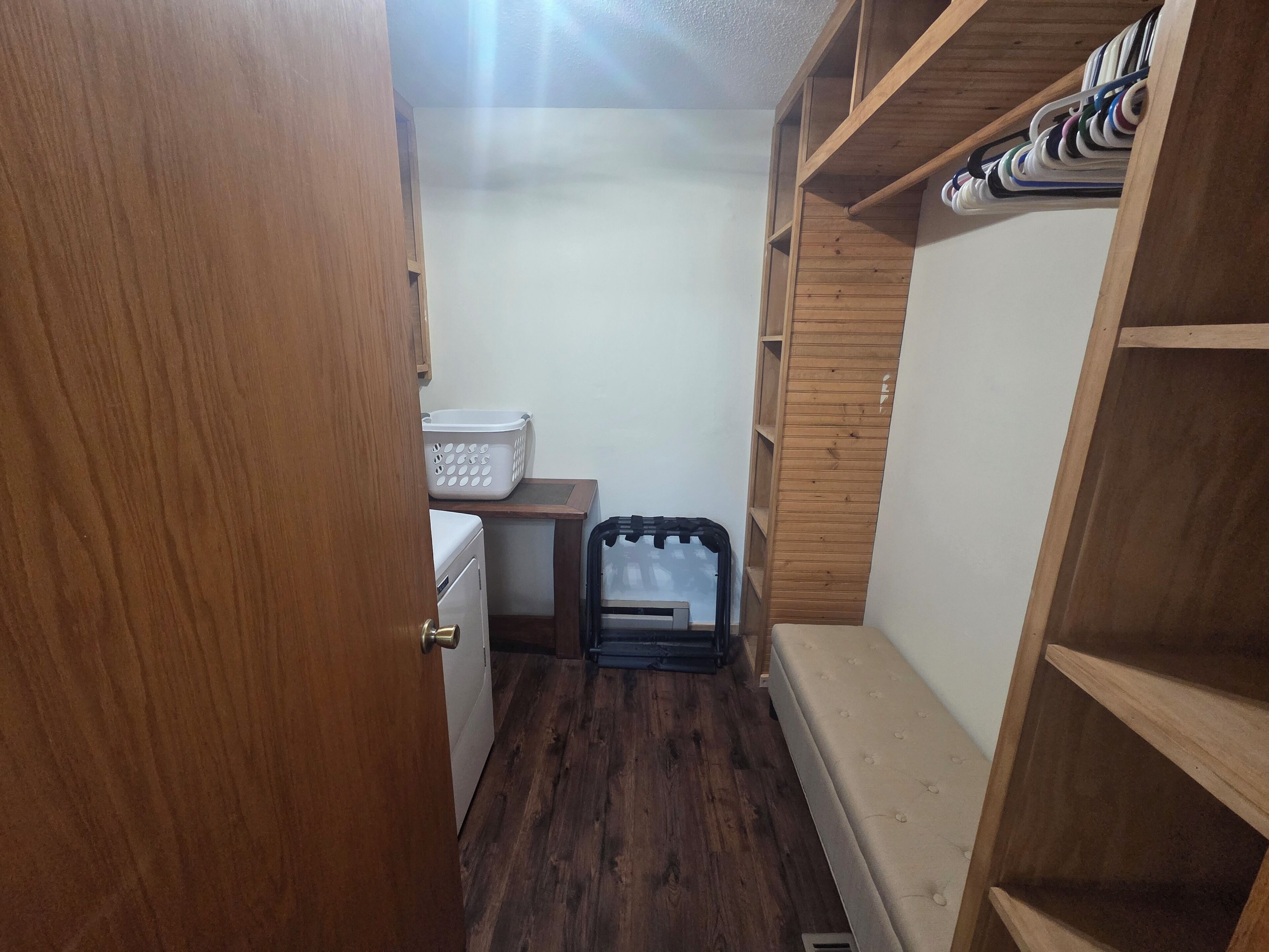 Un couloir étroit au plancher en bois, avec un panier à linge blanc, un meuble de rangement noir et des étagères en bois.