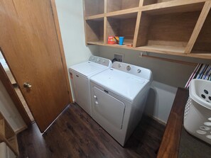 A laundry room with a front-loading washer and dryer, wooden shelving above, and a white laundry basket.