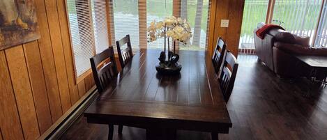 A dining area with a wooden table, chairs, a vase with flowers, and a view through large windows.