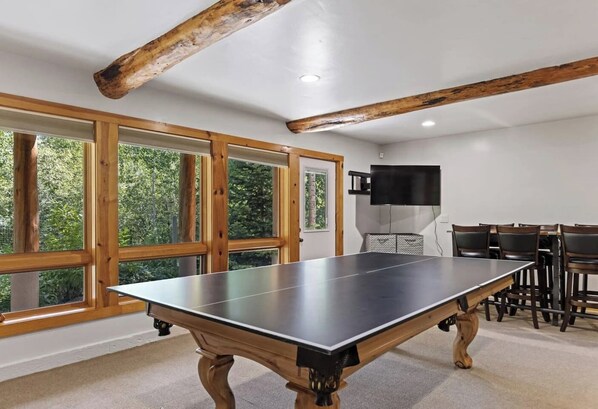 Ein Tischtennis-Tisch in einem Raum mit großen Fenstern, Holzbalken und einem Flachbildfernseher.