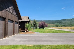 A building with a garage, a driveway, and a grassy area with trees. - Retreat on the Edge of Victor with New Hot Tub (Victor)