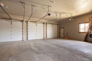 A spacious garage with multiple white garage doors, a concrete floor, and ceiling-mounted lighting fixtures. - Retreat on the Edge of Victor with New Hot Tub (Victor)