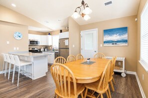 A modern kitchen with white cabinets, a central island, and a dining area with wooden chairs. - Private Pool · 3-Min Walk to Beach · Sleeps 8, Loaded, Pets OK (Nags Head)