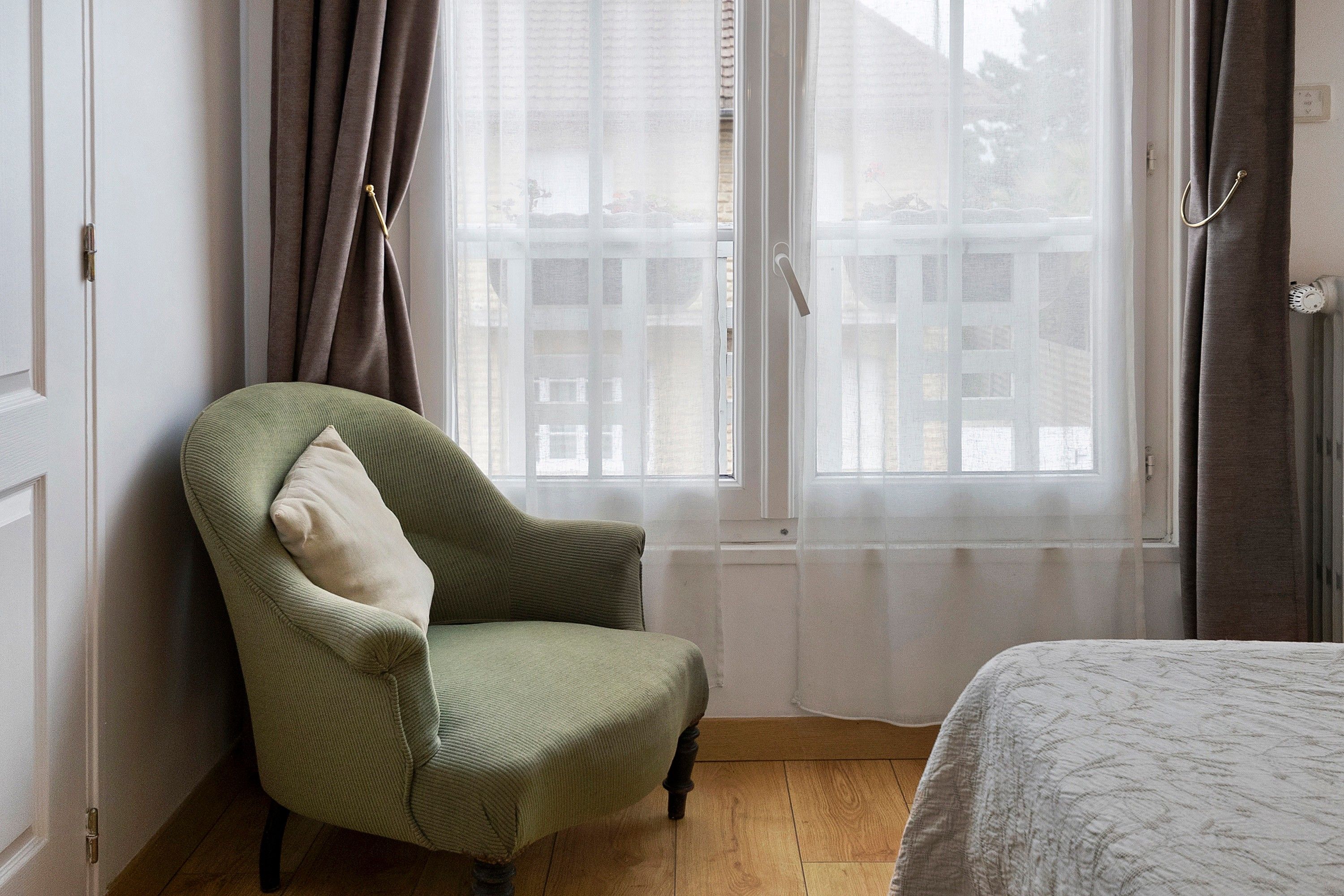 Um quarto com uma poltrona verde, piso de madeira, janela com cortinas e uma cama com colcha estampada.