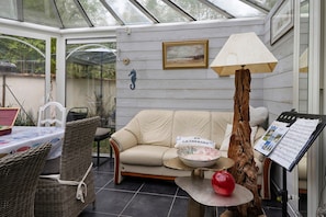 A conservatory with a cushioned seating area, a table with a placemat, a wicker chair, a lamp, and a music stand with sheet music.