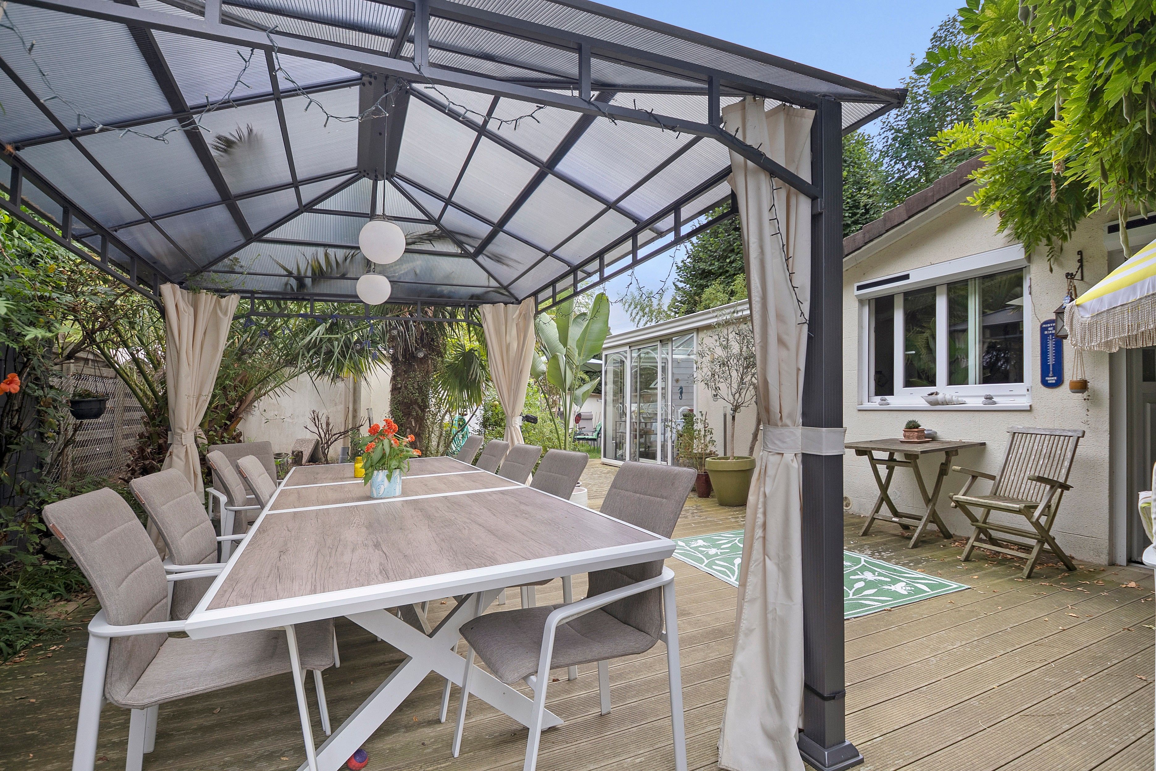 Un espace extérieur abrité, aménagé pour les repas, avec une table et des chaises, entouré de verdure et d’une maison.