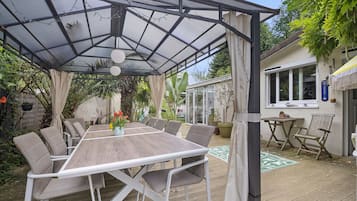 Un espace extérieur abrité, aménagé pour les repas, avec une table et des chaises, entouré de verdure et d’une maison.