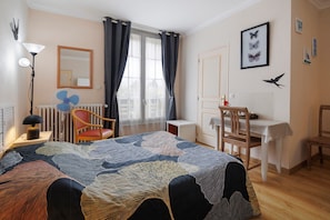 A bedroom with a bed, a desk, a chair, a radiator, a window with curtains, and wall art.