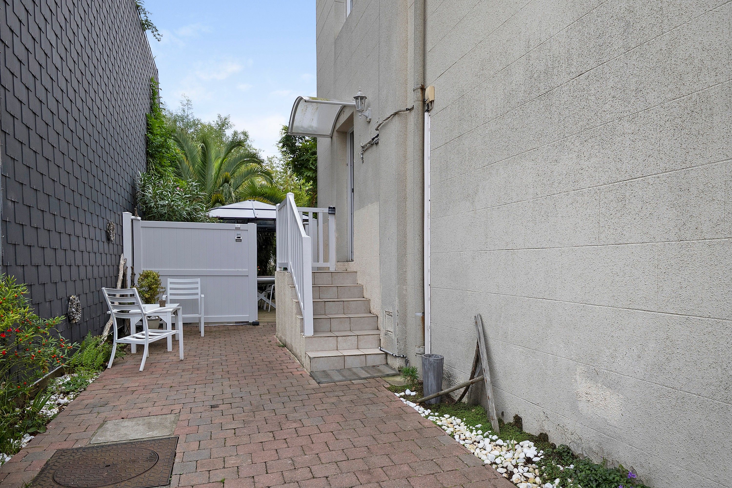 A narrow brick pathway leading to a white door, with a white chair and a small table outside.