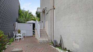 A narrow brick pathway leading to a white door, with a white chair and a small table outside.