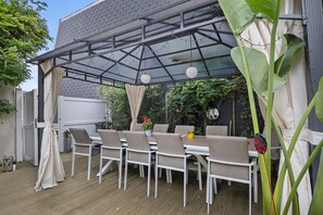 Une terrasse extérieure avec une table et des chaises, une pergola dotée d’un toit en verre et une végétation luxuriante.