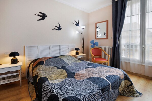 A bedroom with a bed, a chair, a desk, a lamp, a radiator, and a window with curtains.