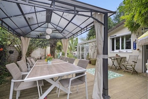 A covered outdoor dining area with a table and chairs, surrounded by greenery and a house.