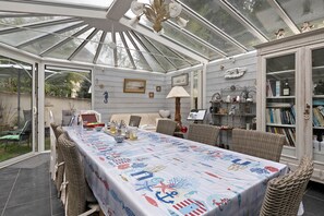 A conservatory with a table set for dining, wicker furniture, and a bookshelf filled with books and decorative items.