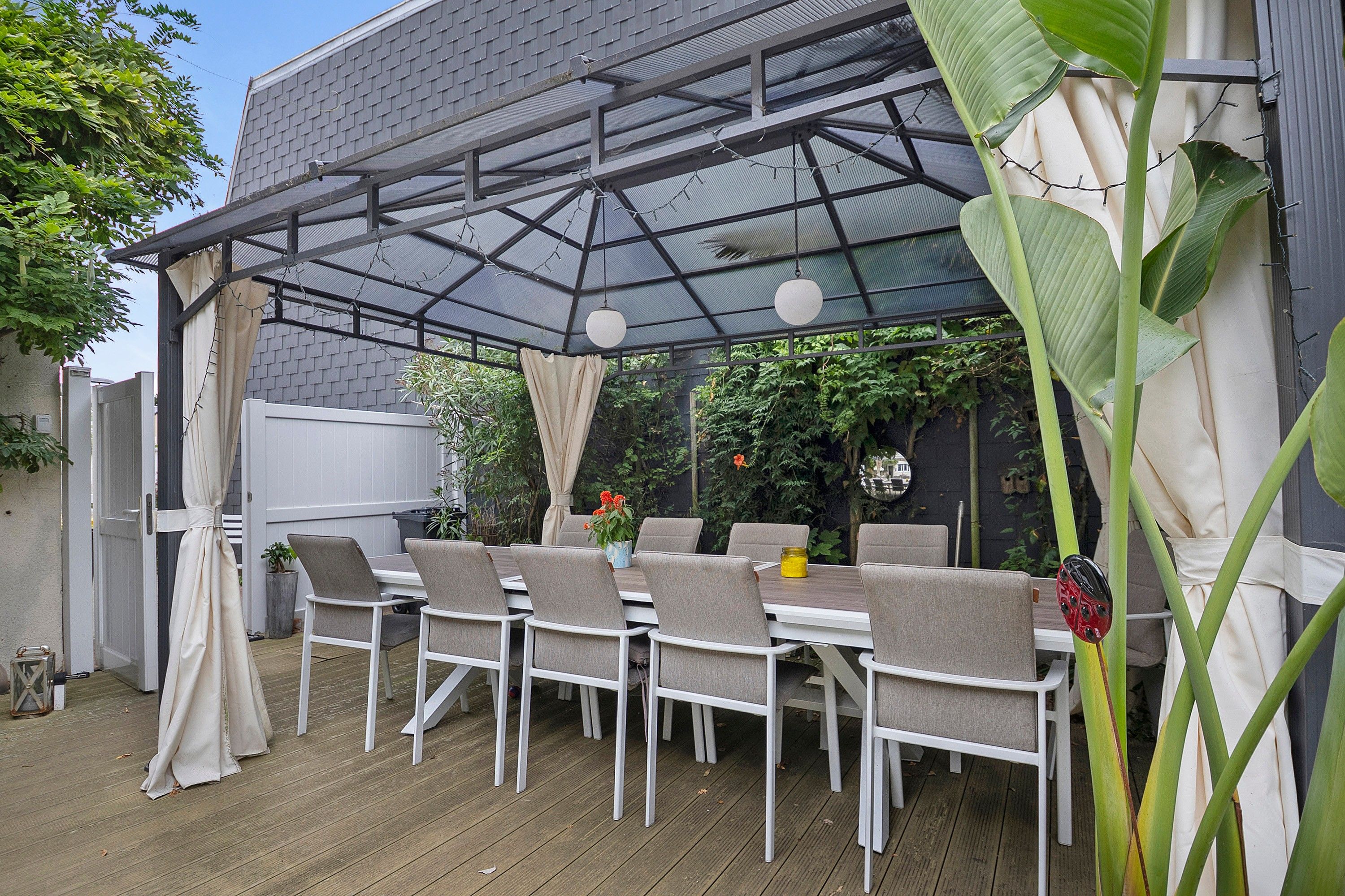 Un patio exterior con una mesa y sillas, una pérgola con techo de vidrio y abundante vegetación.