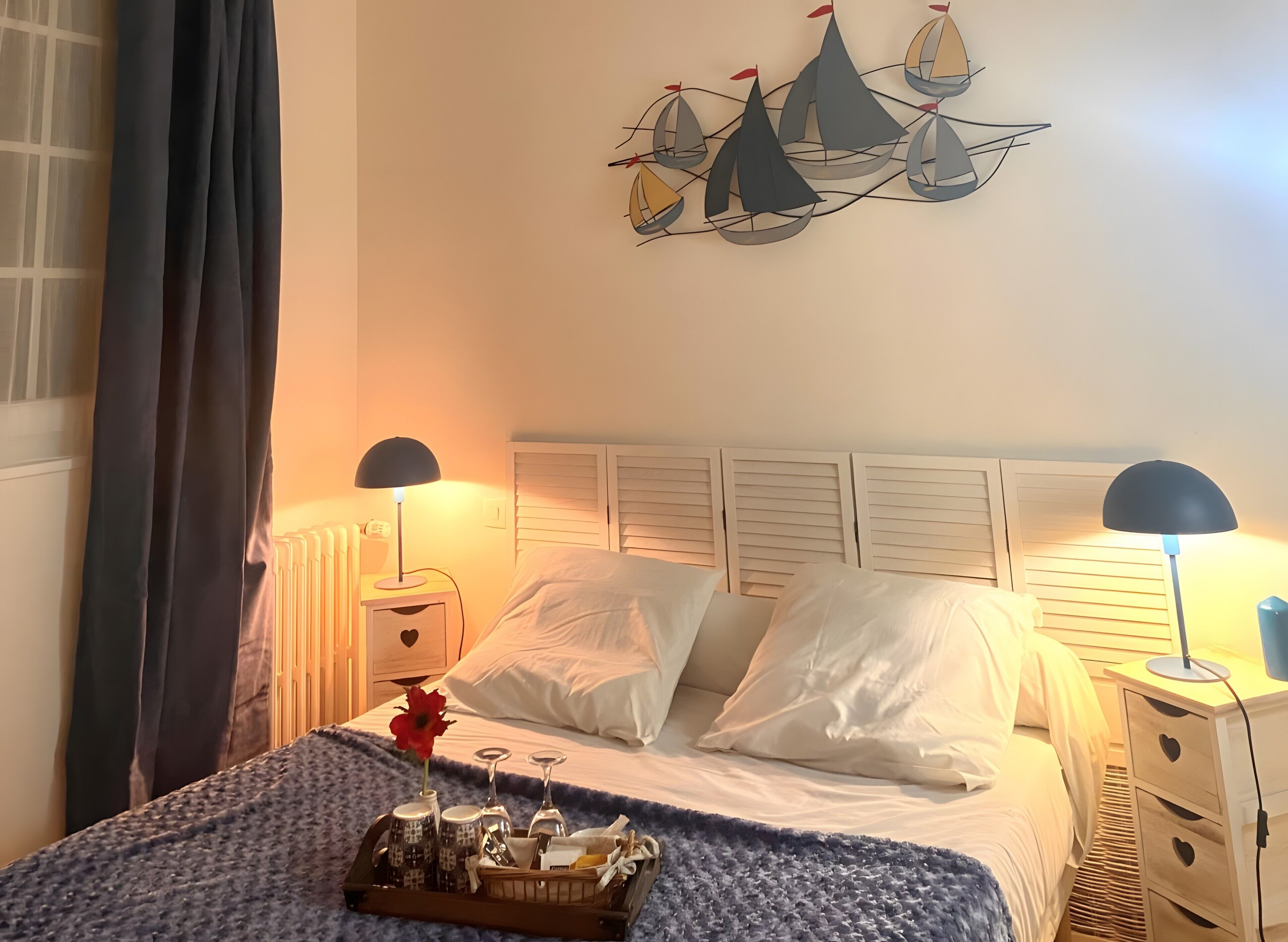 Una cama bien hecha con sábanas blancas, dos lámparas de noche, un cuadro de veleros en la pared y una bandeja con vasos y una flor sobre la cama.