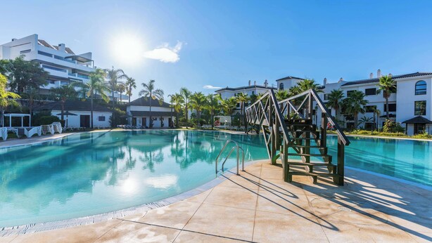 A clear swimming pool with a wooden deck and lounge chairs, surrounded by palm trees and modern buildings. - Mirador Estepona Hills 15 | Pool & Private Jacuzzi (Estepona)