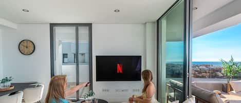 A modern living room with a large flat-screen TV mounted on the wall, a sofa, and a dining area with chairs.