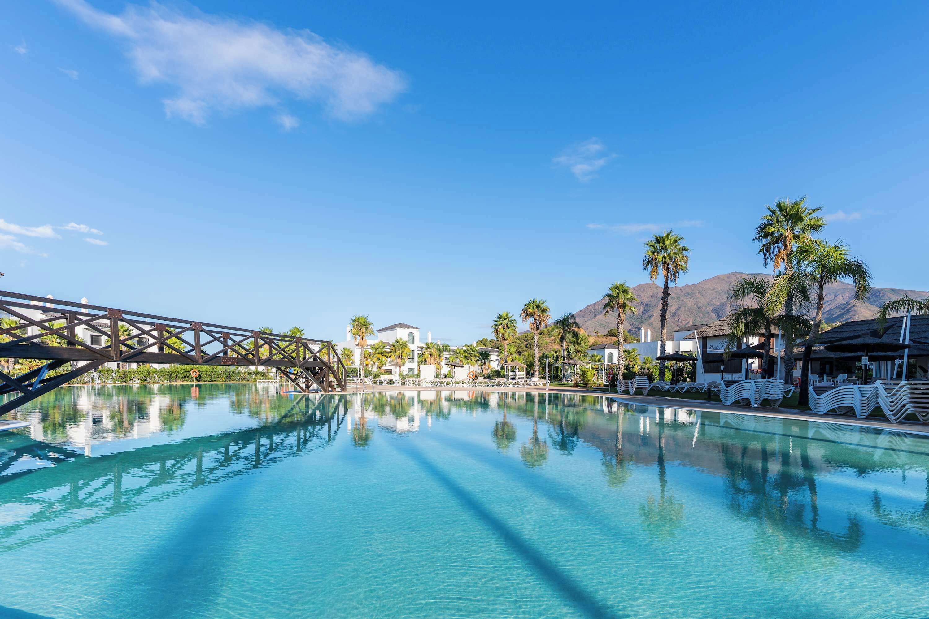 Une piscine aux eaux cristallines, avec un pont, des palmiers et une montagne en arrière-plan.