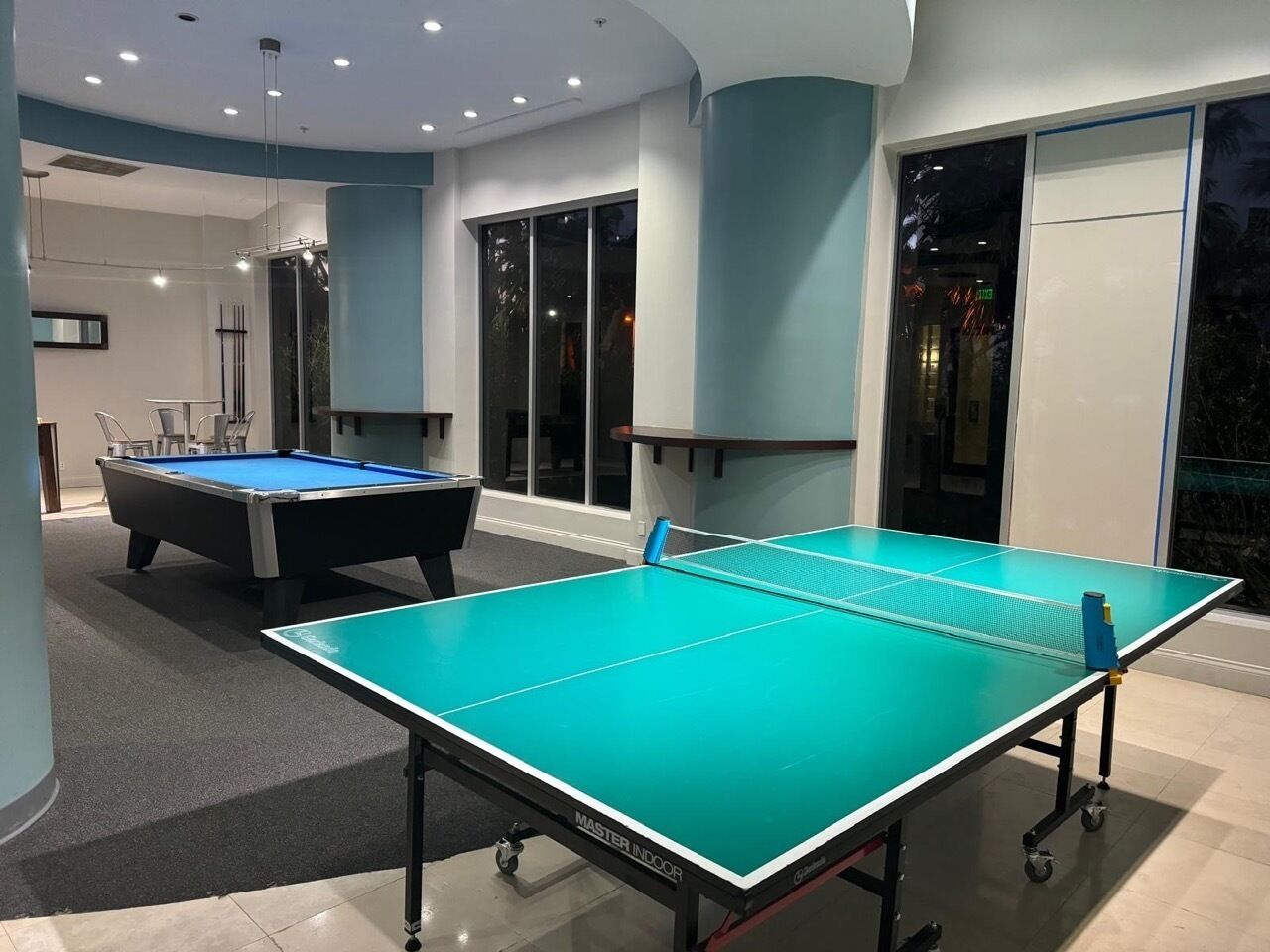 Een pooltafel en een tafeltennistafel in een kamer met een modern design, grote ramen en een vloerbedekking.