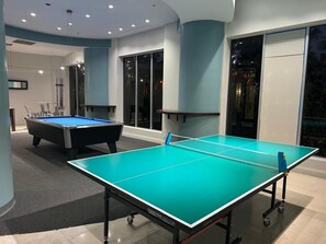 Una mesa de billar y una mesa de ping pong en una sala con diseño moderno, ventanas amplias y suelo alfombrado.