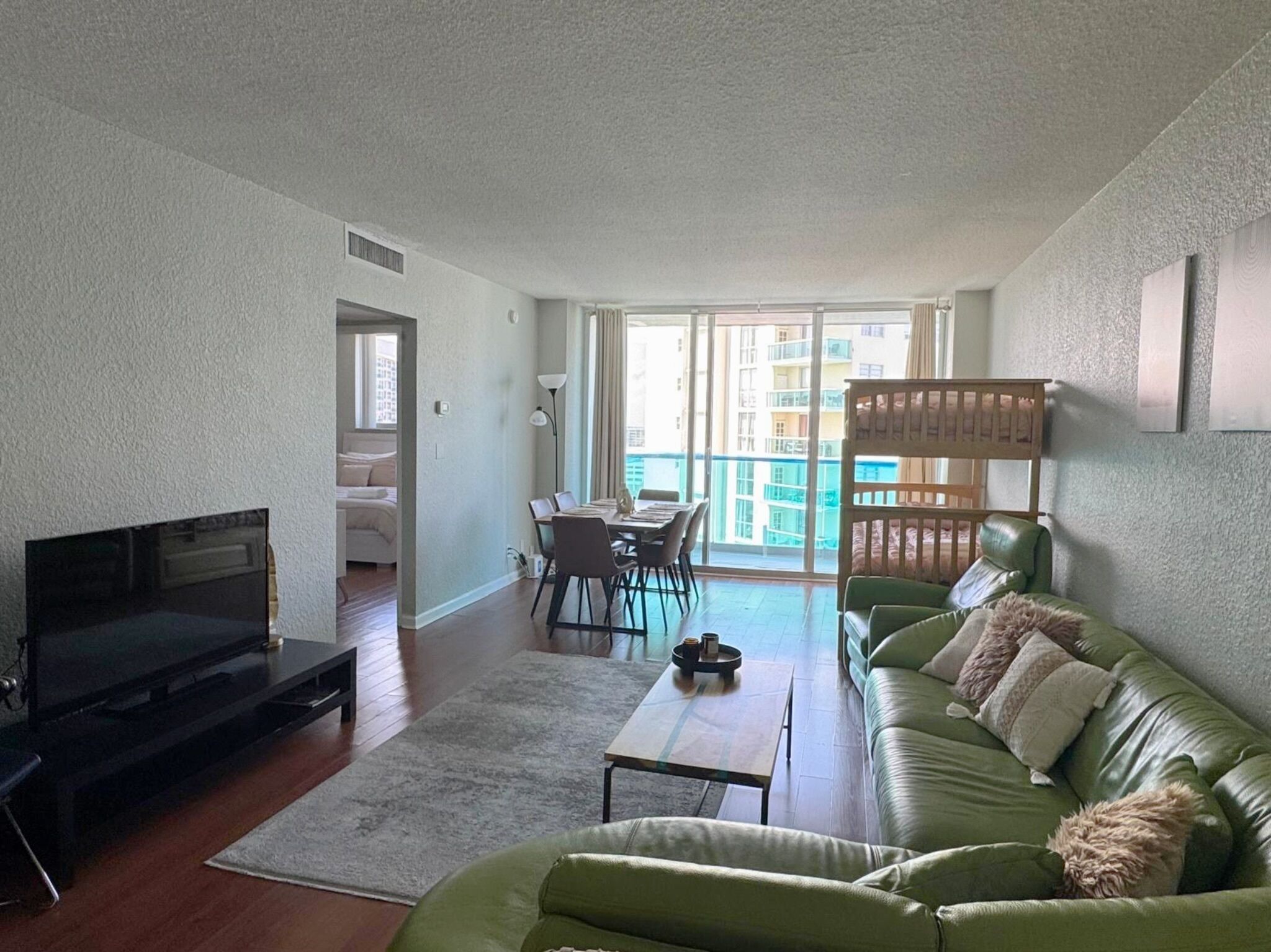 Una sala de estar moderna con un sofá verde, un televisor de pantalla plana, una mesa de comedor con sillas y un balcón con vistas a edificios.
