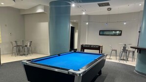 Uma mesa de bilhar em um quarto com uma televisão de tela plana na parede, uma cadeira e uma pequena mesa.
