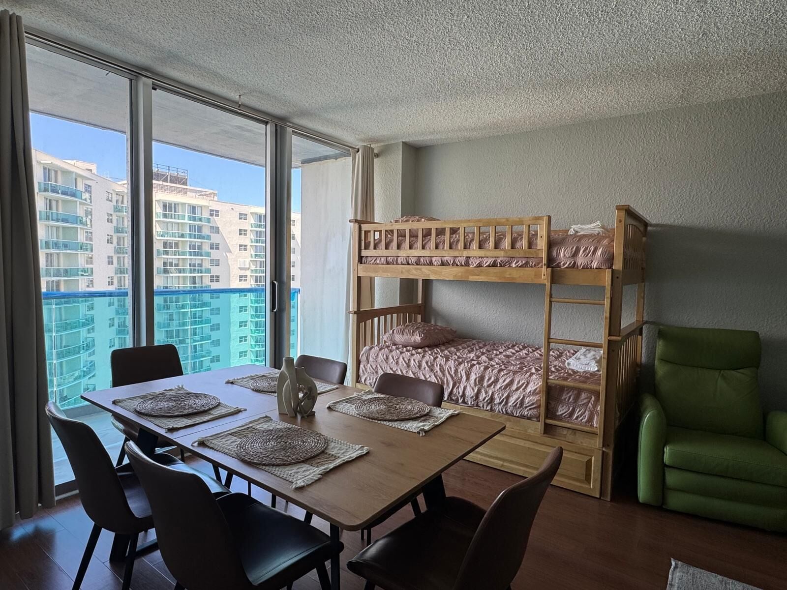 A room with a bunk bed, a dining table with chairs, and a view of high-rise buildings.