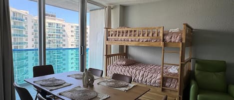 A room with a bunk bed, a dining table with chairs, and a view of high-rise buildings.