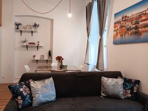 Ein Wohnzimmer mit einem dunklen Sofa, dekorativen Kissen, einem Esstisch mit Stühlen, einer Vase mit Blumen und einem Stadtbild.