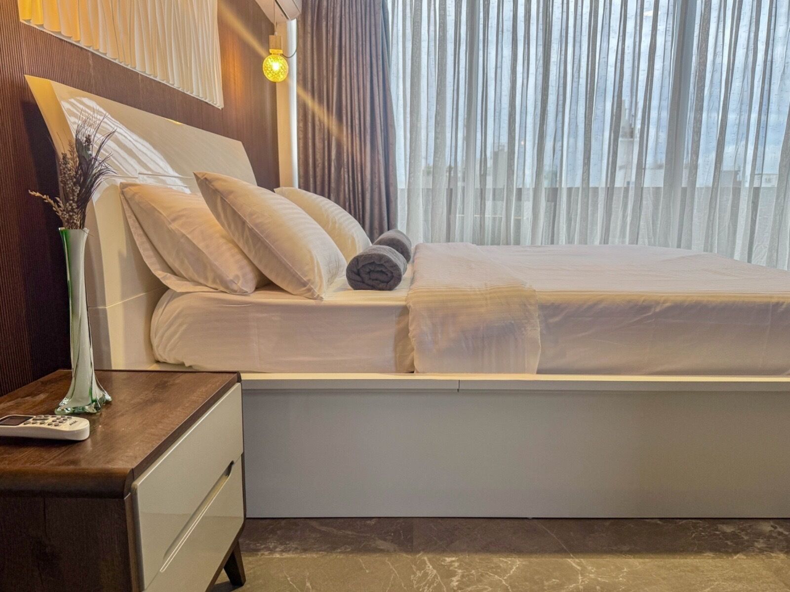 A hotel room with a bed, a nightstand with a lamp, and a vase with dried plants.