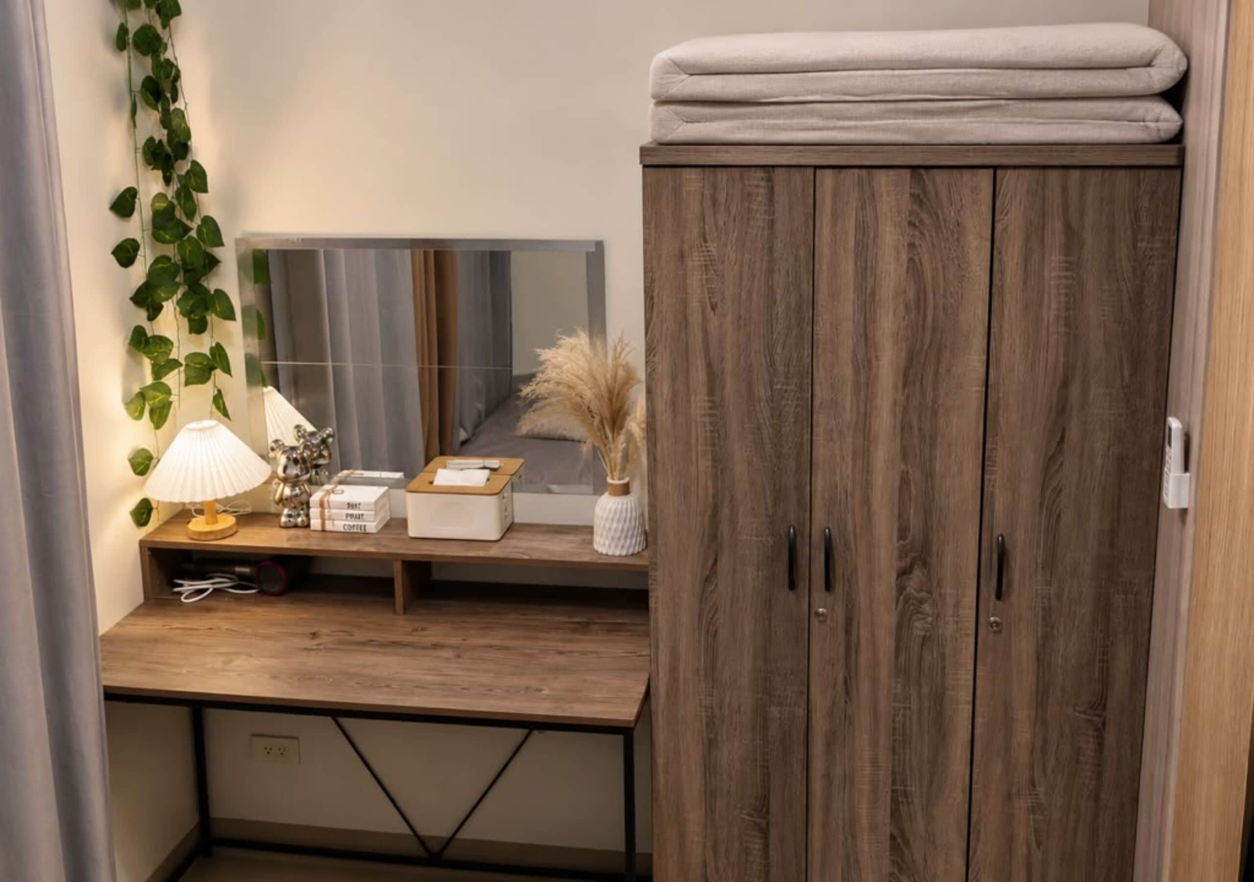 A bedroom with a wooden wardrobe, a bedside table with a lamp, a mirror, and a plant.