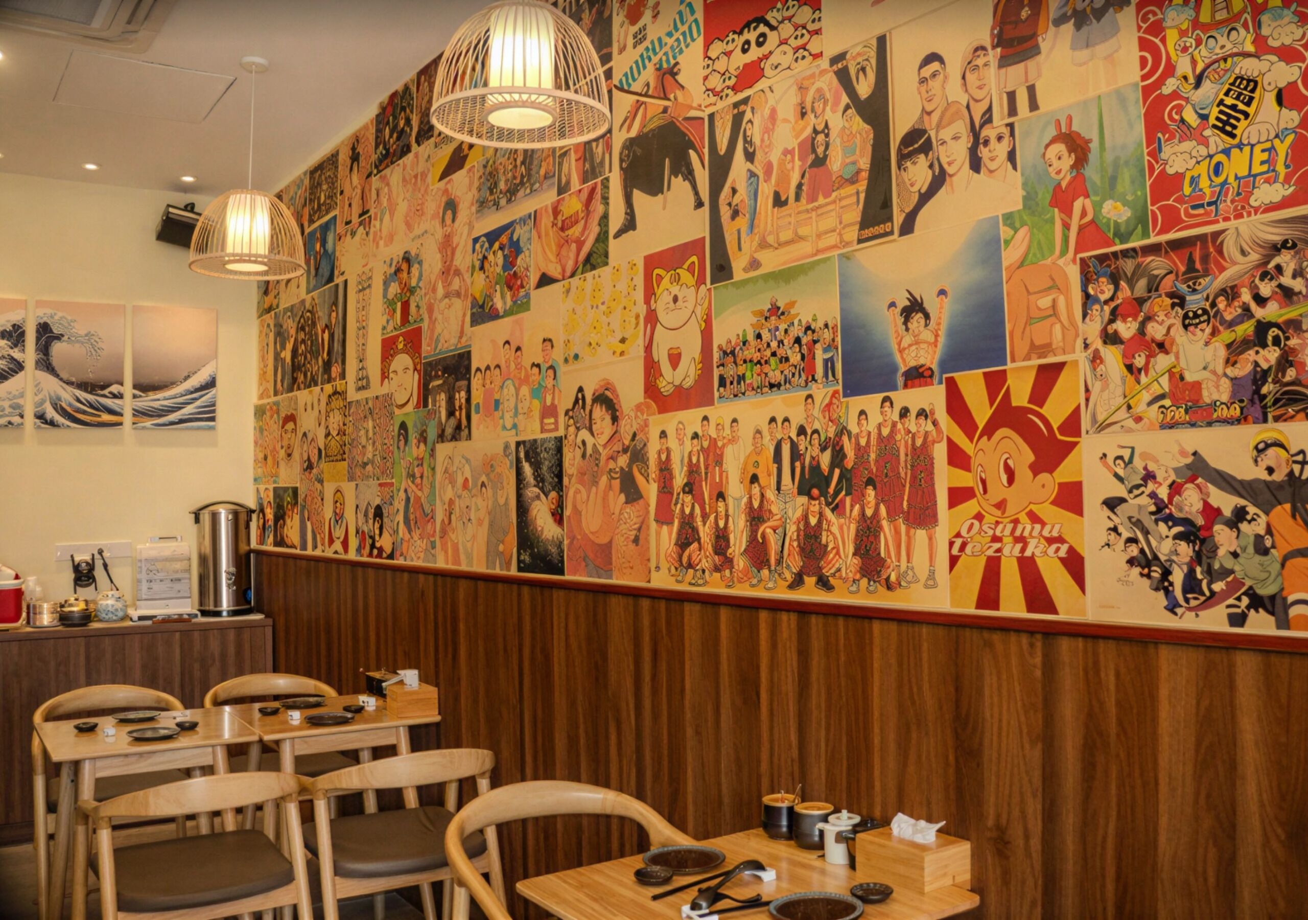 Ein Restaurantinnenraum mit einer Wand, die mit verschiedenen Anime- und Manga-Postern, Holztischen und hängenden Lampen dekoriert ist.