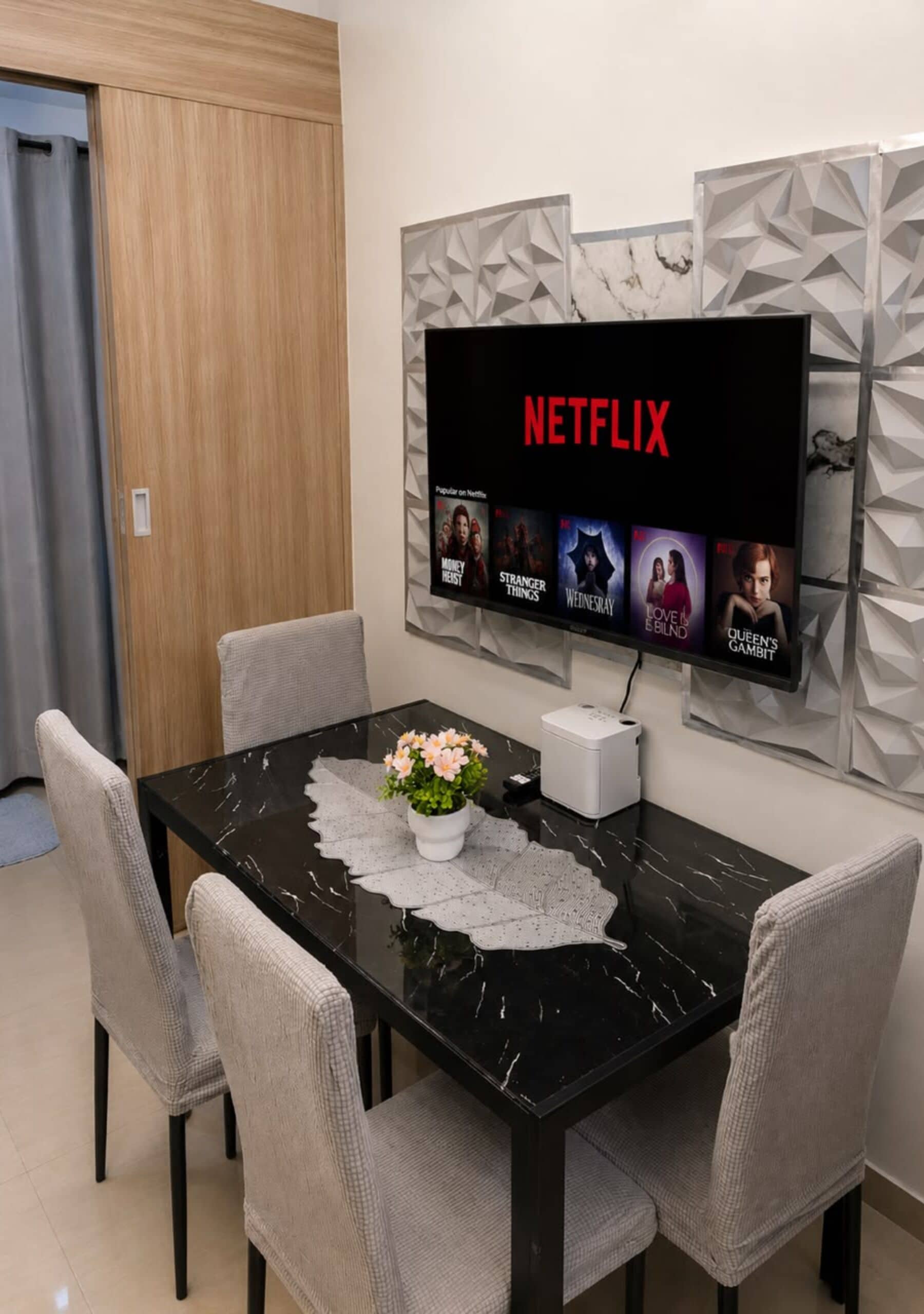 A modern dining area with a black marble table, four grey chairs, a white vase with flowers, and a large flat-screen TV mounted on a wall displaying Netflix content.