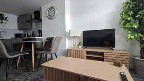 A modern living room with a flat-screen TV, a dining area with chairs, a kitchen with cabinets, and a potted plant.