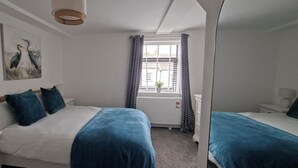A bedroom with two beds, a nightstand, a bedside table, a mirror, a window with curtains, and a painting on the wall.