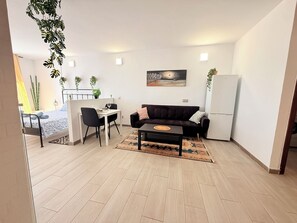 Una sala de estar moderna con un sofá, una mesa de comedor y sillas. Hay un refrigerador, un cuadro en la pared y una planta en maceta.