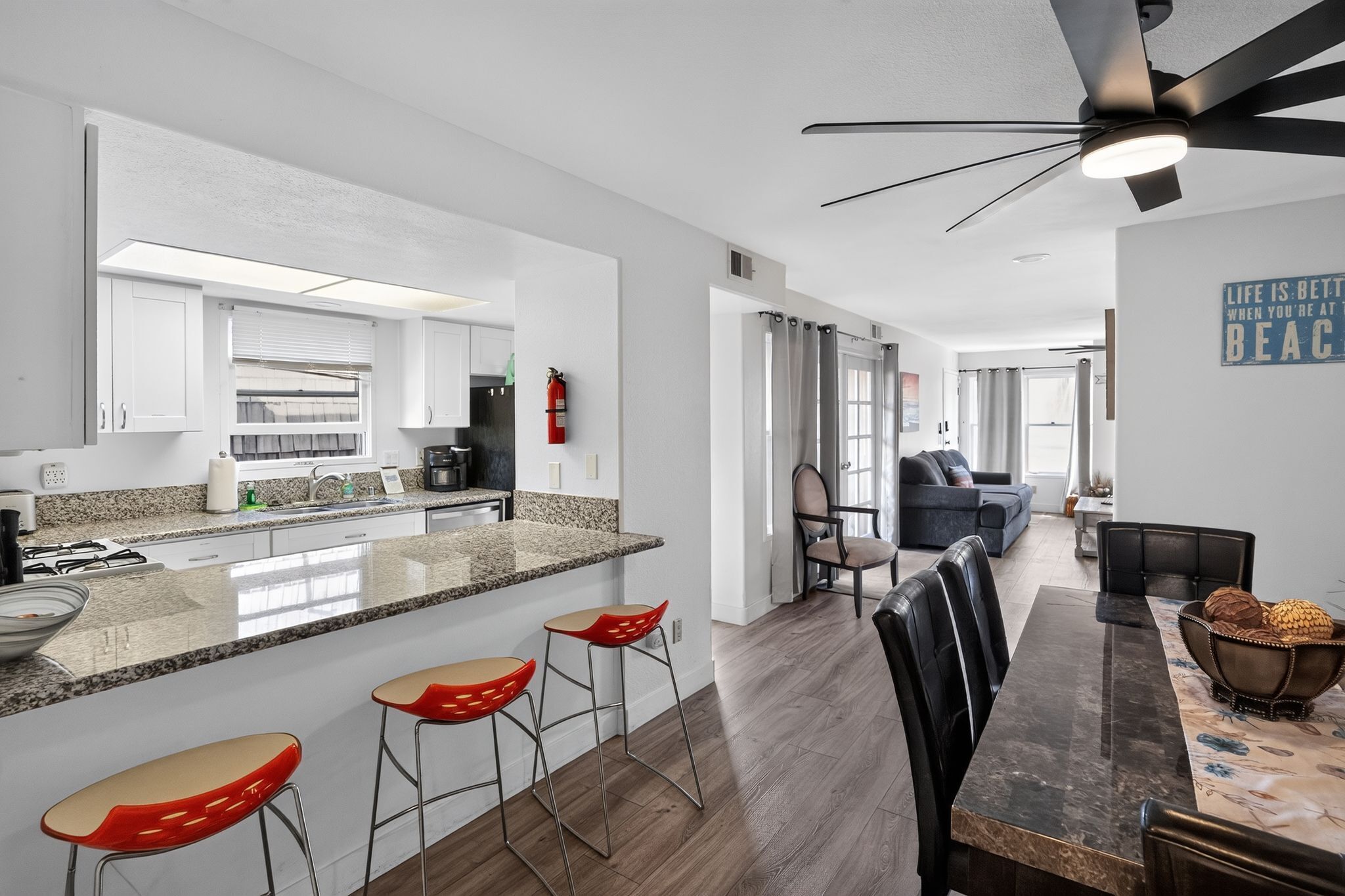 A modern kitchen with granite countertops, a dining area with a wooden table and bar stools, and a living room with a sofa and a coffee table.