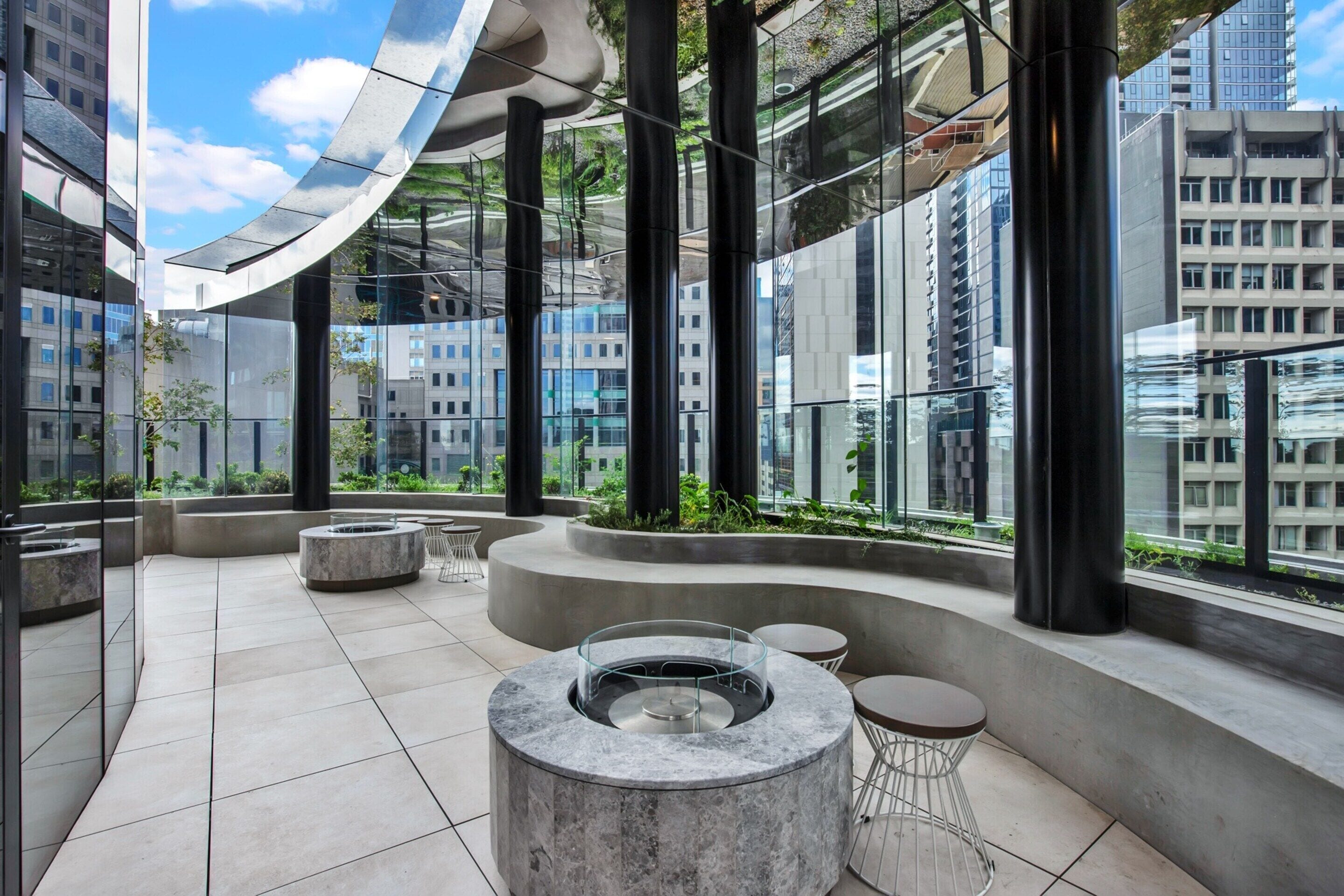 A modern outdoor lounge area with a circular seating arrangement, large windows, and a view of surrounding buildings.