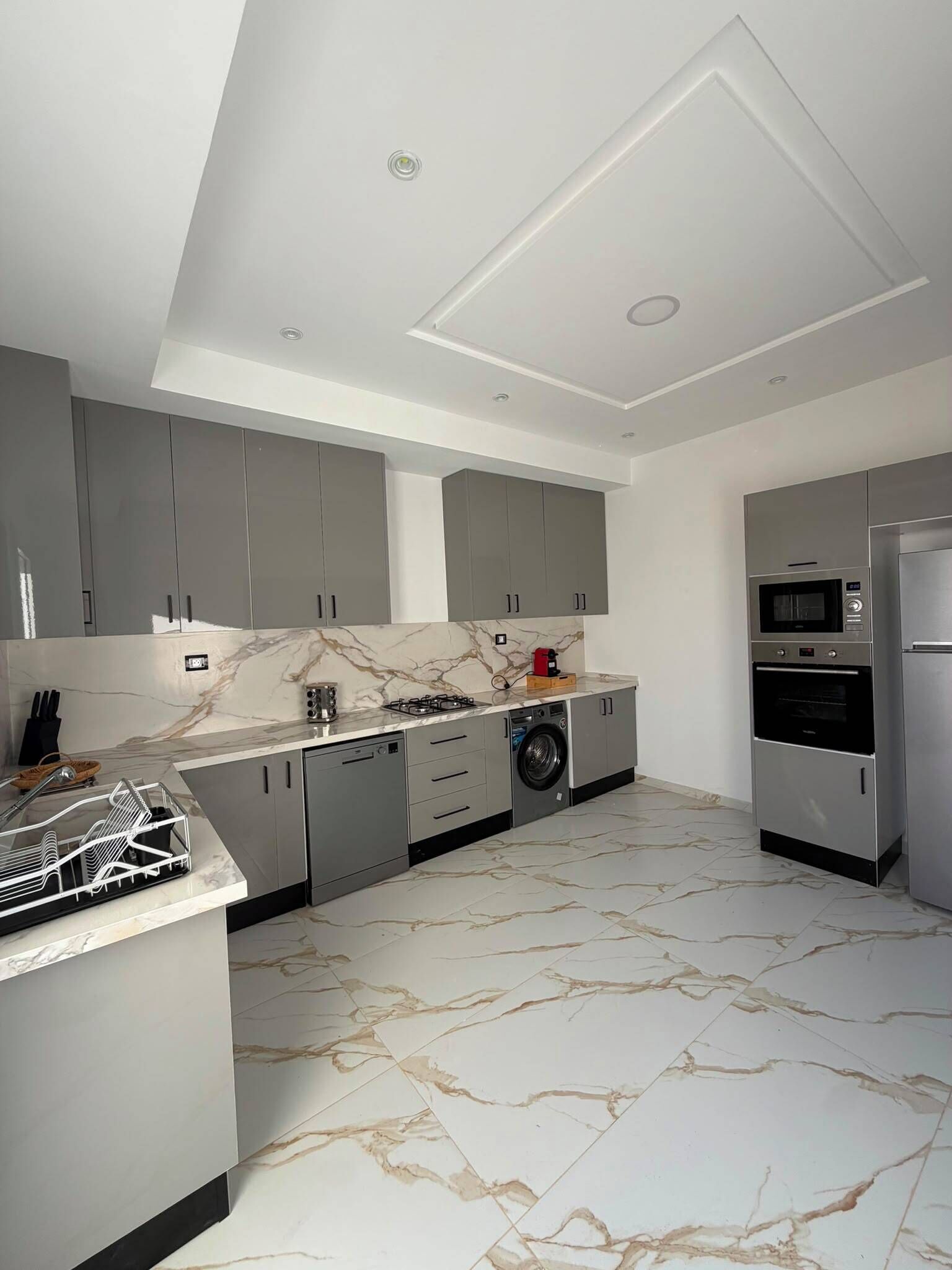 A modern kitchen with grey cabinets, a built-in dishwasher, a microwave, and a stainless steel refrigerator.