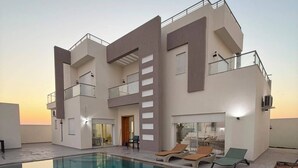 A modern multi-story building with a rooftop pool area, including lounge chairs and a glass-enclosed dining area. - Villa Younes (Djerba Midun)