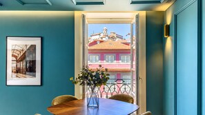 Ein Zimmer mit Esstisch, Stühlen, einer Vase mit Blumen, einem gerahmten Bild an der Wand und Blick auf ein rosafarbenes Gebäude durch eine offene Tür.