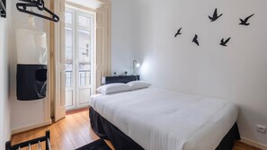 A bedroom with a bed, a hanging bag, a nightstand with a lamp, and a wall with bird silhouettes. - Arroios City Base | 2BR & Workspace by LovelyStay (Lisboa)