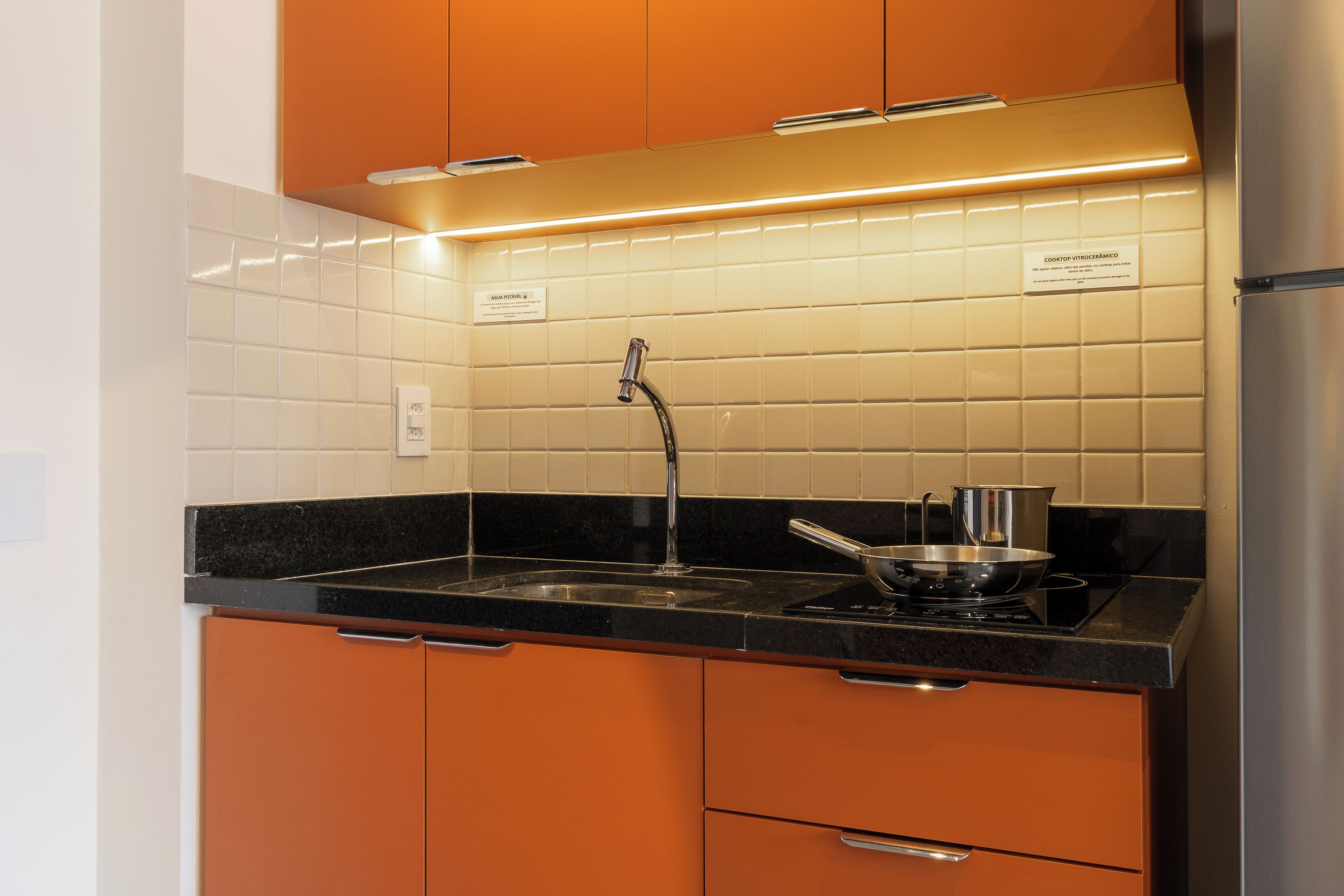 Cozinha com armários laranja, geladeira de aço inoxidável, bancada preta e painel de azulejos.