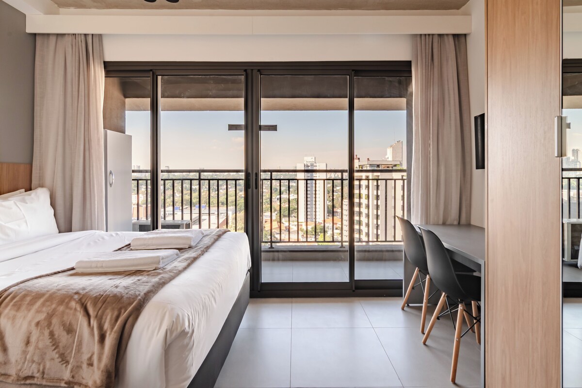A hotel room with a large bed, a desk, and a balcony with a city view.