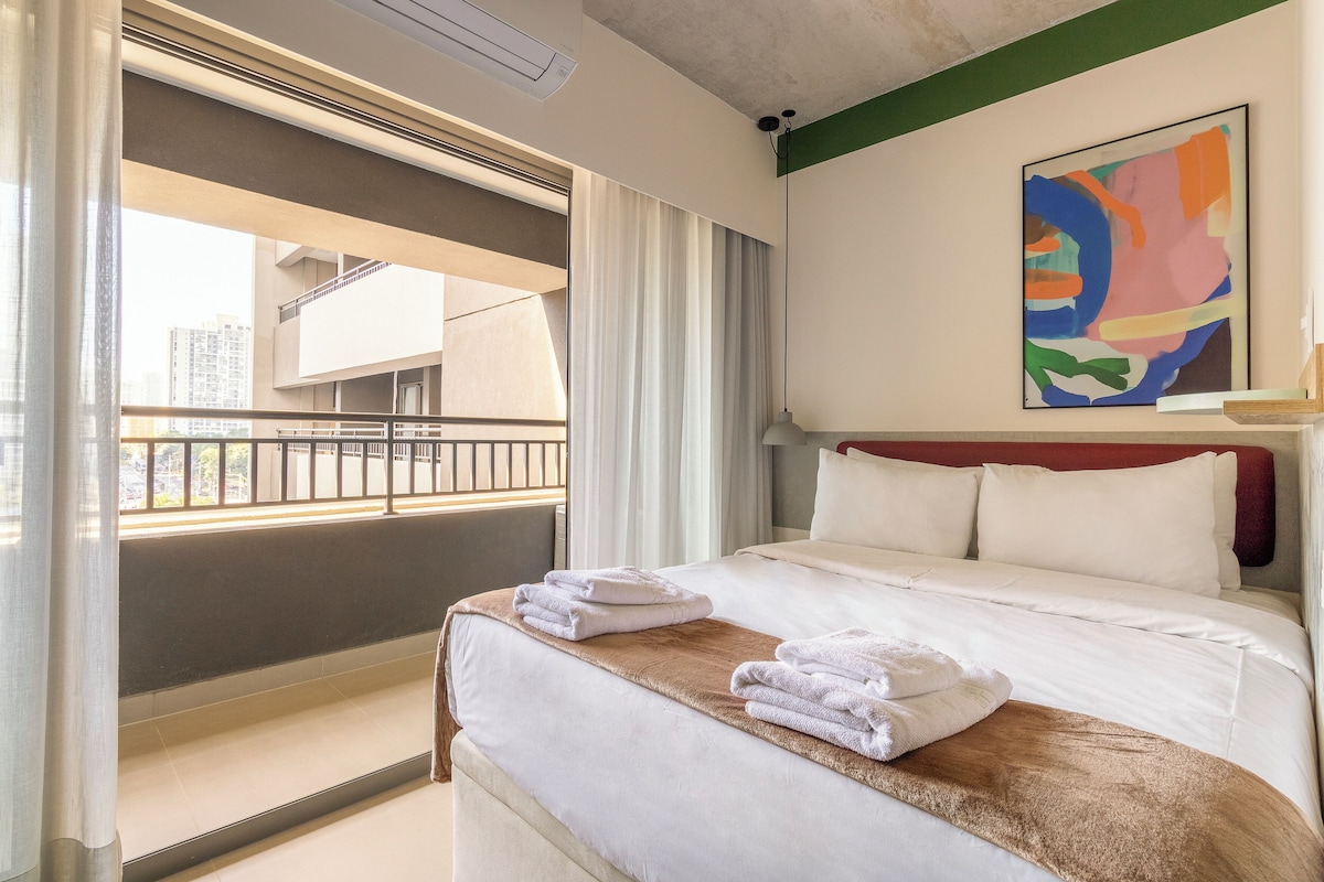 A hotel room with a bed, a view of the outside balcony, and a painting on the wall.